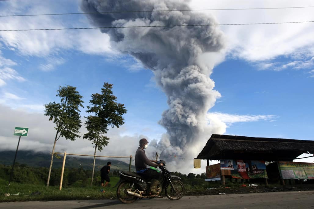 Indonezijos gyventojas važiuoja motociklu pro Sinabungo ugnikalnį, iš kurio kraterio kyla dūmų ir pelenų stulpas. Nuotr. EPA-ELTA